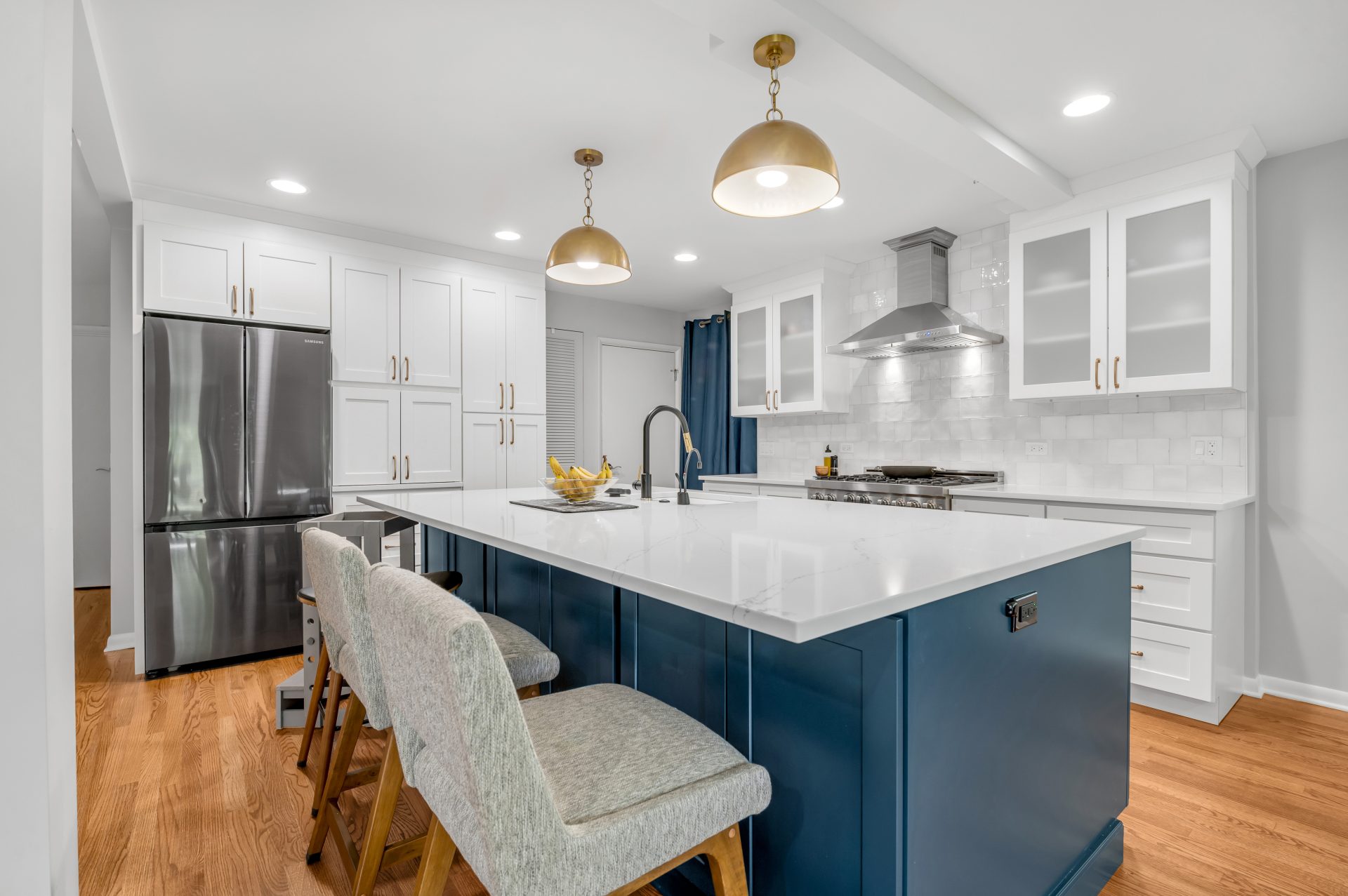 Remodeled kitchen with white square backsplash tiles, white quartz countertop, stainless-steel appliances, exposed stainless steel hood, white and blue two tone cabinets, brass kitchen hardware, two brass kitchen pendants, island sink, and hardwood floors