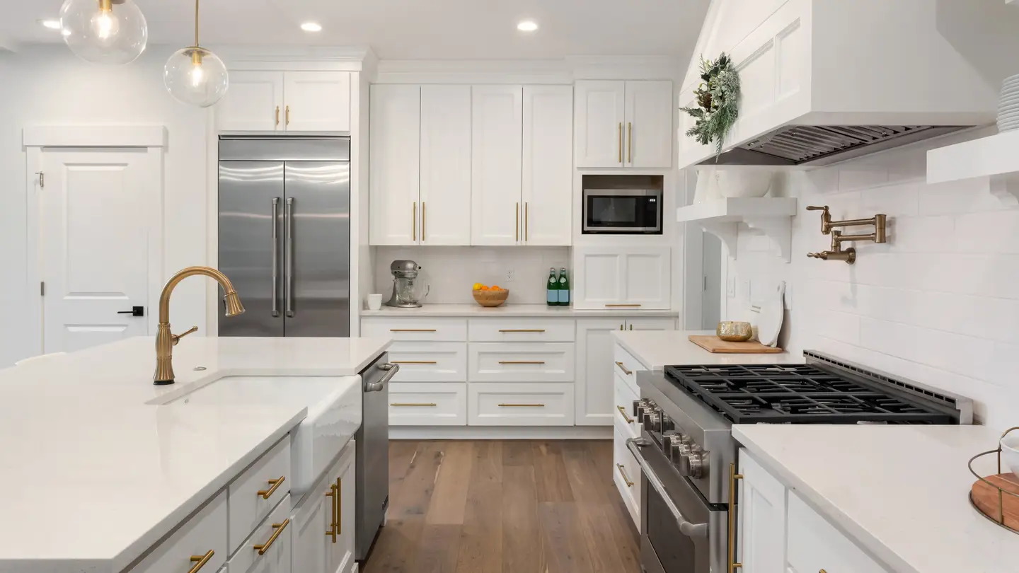 All-white kitchen with silver appliances and a gold sink faucet on a white island.