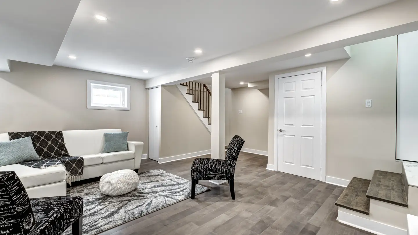 Finished basement with gray flooring and light gray walls. The basement has a zebra-patterned chair, a gray and white rug, and a white couch under a window.