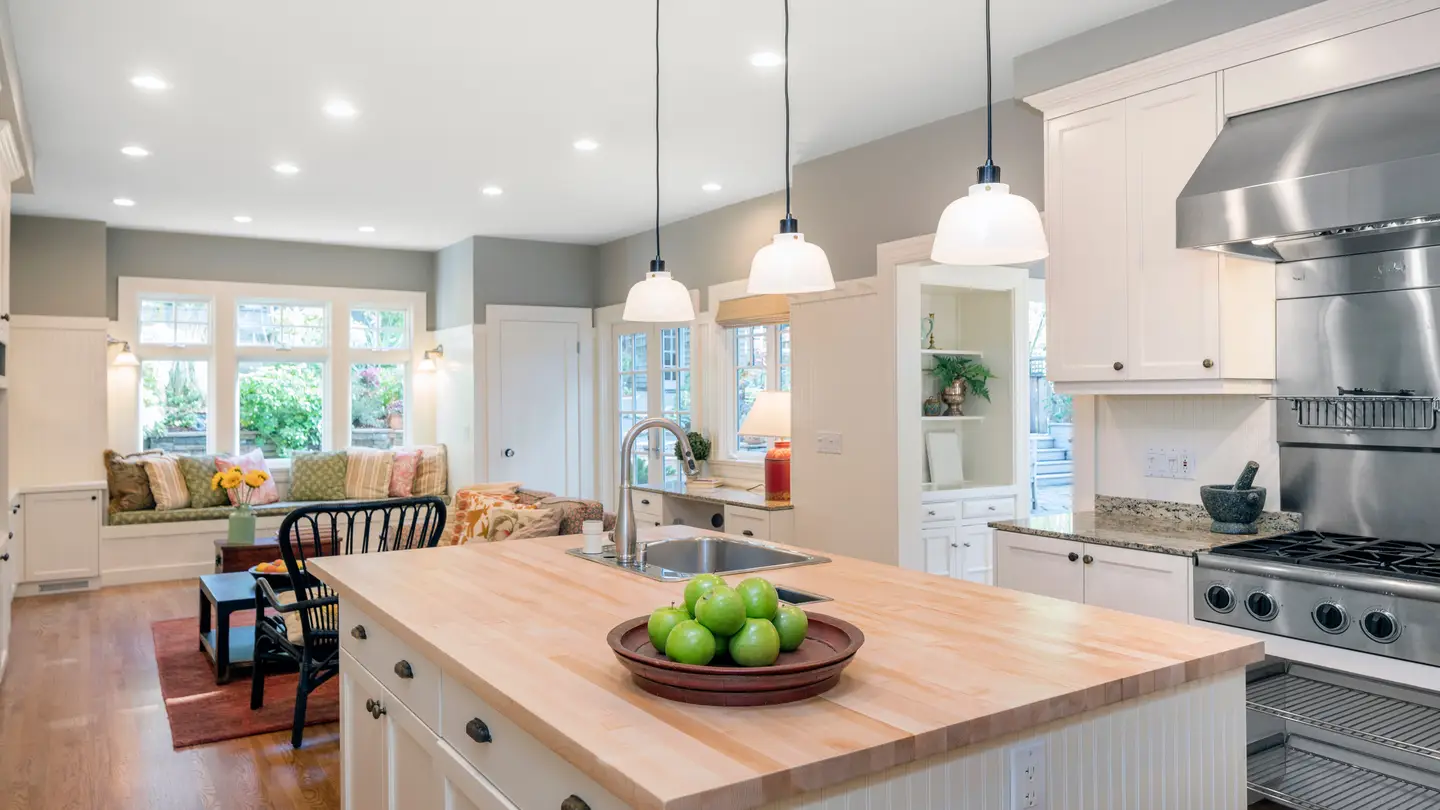 Large white kitchen with an island with brown cutting board top and light fixtures hanging overhead.