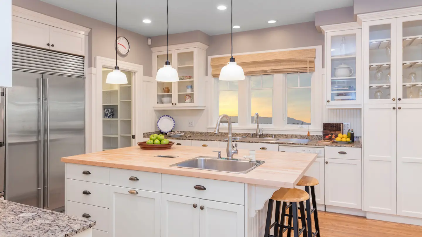 Kitchen with an island that has white cabinets and a cutting board countertop with surrounding white cabinetry and light fixtures hanging over the island.