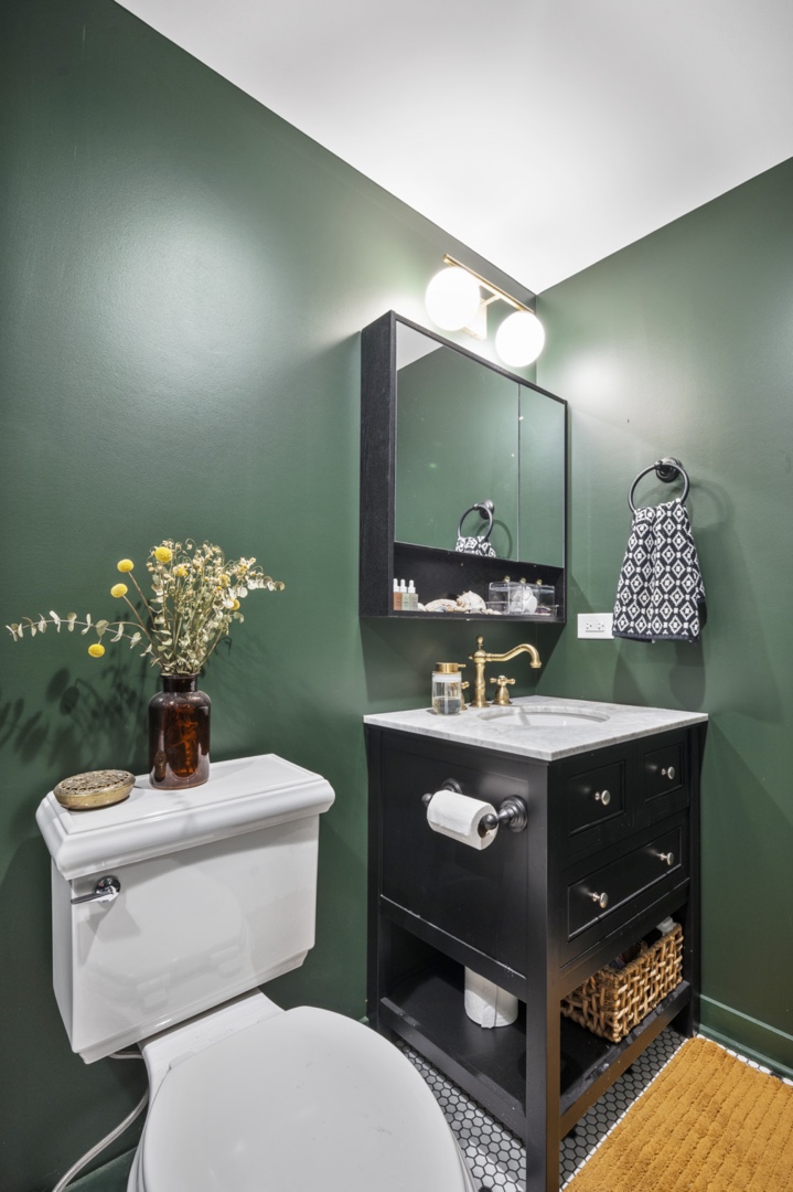 A bathroom with a white toilet, dark green walls, vanity lights and a white sink with simple decor.