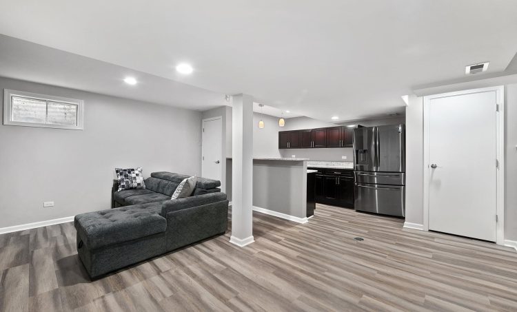 A spacious basement with white walls and gray accents with an attached kitchen and stainless steel appliances