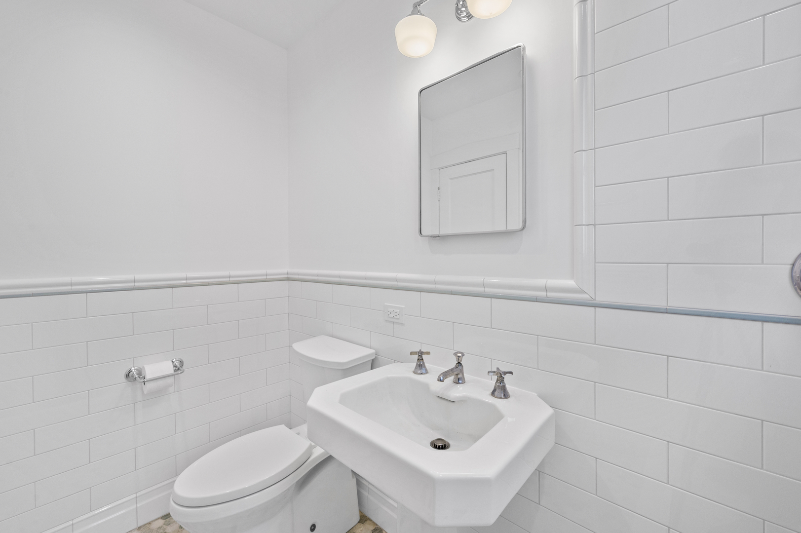 An all-white bathroom with subway tile walls, a white toilet, and sink with a mirror and vanity lights.