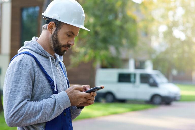 Young male builder in hardhat using smartphone, standing outdoors while working at cottage construction site. Profession, construction and building concept. Side view