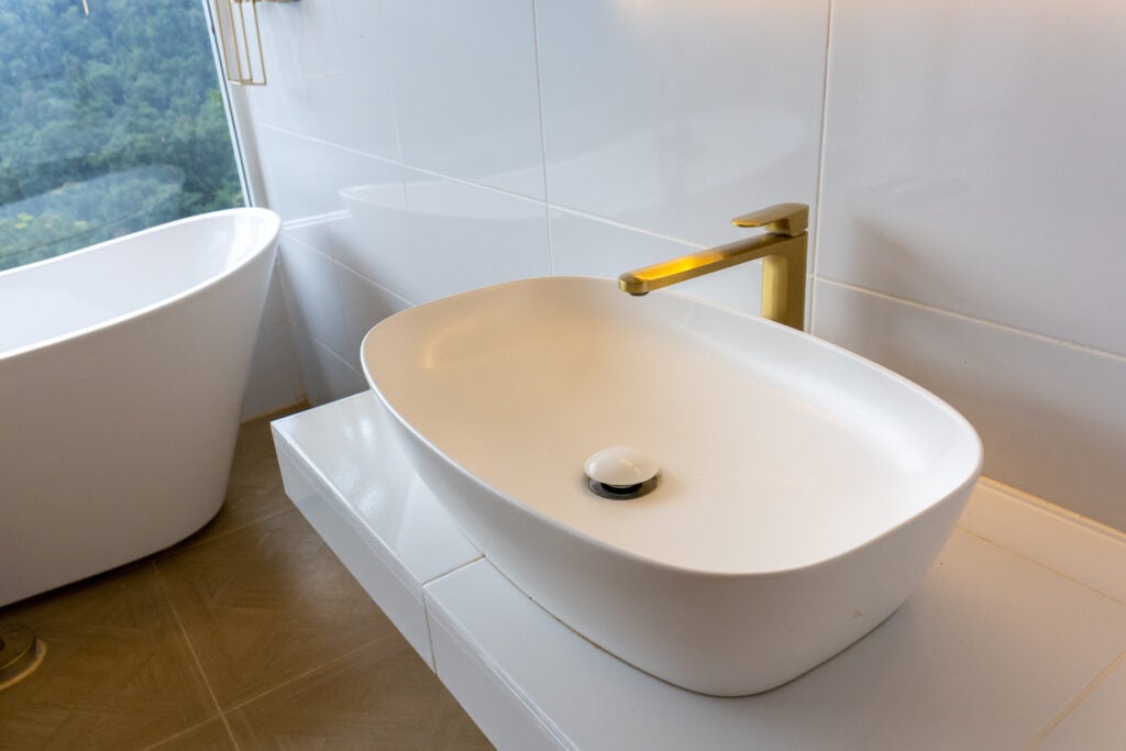 single bathroom vanity with sink A modern white oval bathroom sink with gold faucet on floating vanity overlooking forest view, minimalist clean design and soft natural light creating calm ambiance