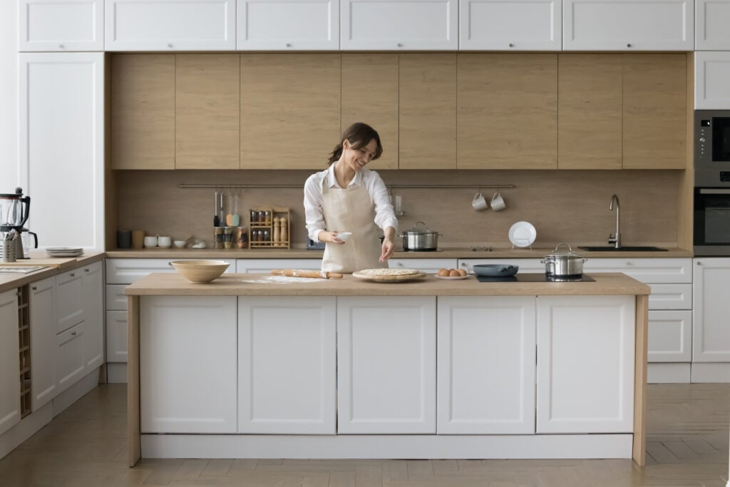 Happy young baker girl preparing pizza, pie, dropping grated cheese on rolled dough, baking in home kitchen modern interior, enjoying culinary hobby, cooking activity, smiling